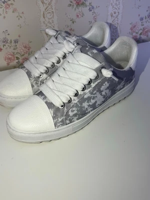 Gråvita sneakers från NUMERIS - Snygga sneakers från NUMERIS  grått och vitt med coolt camomönster. Skorna har vita snören, tåparti och sula i syntetmaterial samt textilfoder. Perfekta för dig som gillar streetstyle och vill sticka ut lite extra.