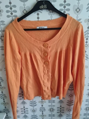 Persikofärgad kofta från Gina Tricot - Söt persikofärgad kofta från Gina Tricot med rund halsringning och tre stora knappar framtill. Koftan har långa ärmar, ribbade muddar och ett lätt rynkat parti över bysten för en avslappnad look.