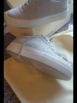 NYA sneakers från Axel Arigato i mocka - NYA snygga sneakers från Axel Arigato med klassisk låg profil och tjock vit sula. Färg light grey. Skorna är tillverkade i mocka och har diskreta detaljer samt ton-i-ton snörning. Perfekt för dig som gillar stilrena och trendiga skor.Endast använda inomhus ett fåtal tillfället. Finns kartong, påsar och kvitto. 