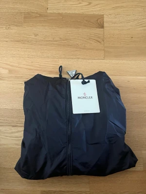 Marinblå Moncler vindjacka med huva - Snygg marinblå  vindjacka från Moncler med huva och dragkedja framtill. Jackan har två fickor med knappar och Moncler-logga på ärmen. Materialet är lätt och vattenavvisande, perfekt för blåsiga dagar. Huva med röd och vit detalj upptill. Passar m om du är runt 180 annars passar den s