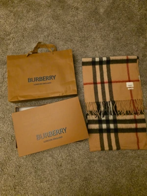 Beige rutig halsduk från Burberry - Säljer en klassisk beige halsduk från Burberry med ikoniskt rutigt mönster i svart, rött och vitt. Halsduken har fransar i ändarna och är tillverkad i mjukt material som känns lyxigt och tidlöst. Perfekt för att lyfta din outfit.