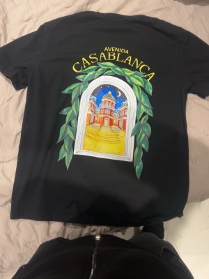 Svart Casablanca t-shirt med tryck - Svart t-shirt från Casablanca med färgglatt tryck av ett fönster med grönska och byggnad på både fram- och baksida. Texten 'Avenida Casablanca' i gult ovanför motivet. Rund hals och korta ärmar. Perfekt för dig som gillar unika prints och streetwear.