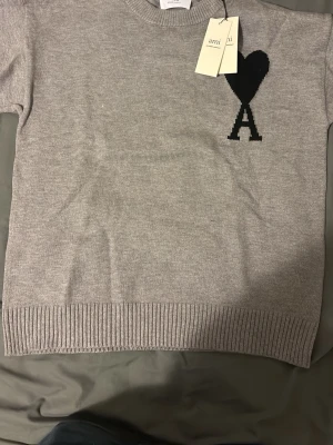 Grå stickad tröja från Ami Paris - Snygg grå stickad tröja från Ami Paris med svart hjärta och bokstaven A broderat på bröstet. Klassisk rund halsringning och ribbade muddar vid ärmslut och nederkant. Perfekt för en stilren och trendig look.