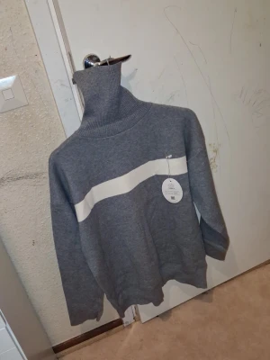 Grå polotröja med vit rand (Man) - Säljer en snygg grå stickad polotröja från FILA med en vit horisontell rand över bröstet. Tröjan har lång ärm, ribbstickad polokrage och muddar. Perfekt för dig som gillar sportiga och stilrena plagg.
