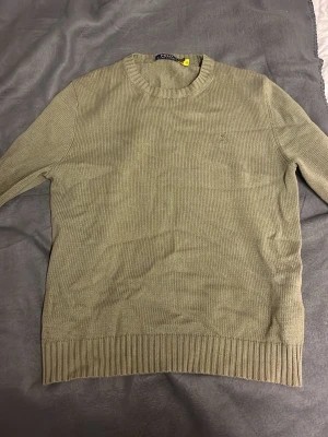 Olivgrön stickad tröja Polo Ralph Lauren - Snygg olivgrön stickad tröja från Polo Ralph Lauren med diskret broderad logga på bröstet. Tröjan har rund halsringning, långa ärmar och ribbade muddar vid ärmslut och nederkant. Perfekt för lager på lager och enkel att matcha. Aldrig använd