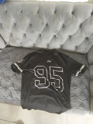 Svart t-shirt med tryck från New Garments - Svart t-shirt från New Garments med stort vitt 95-tryck på framsidan och vita ränder på ärmarna. Klassisk passform och sportig look, tillverkad i 100% polyester. Perfekt för dig som gillar streetwear.