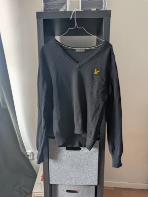 Svart stickad tröja Lyle & Scott - Svart stickad tröja från Lyle & Scott med V-ringning och gul broderad logga på bröstet. Tröjan har ribbade muddar och klassisk passform. Perfekt för lager-på-lager och enkel att matcha med jeans eller chinos.