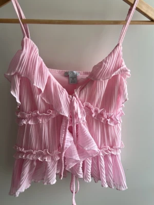 Rosa volangblus med knyt  - Supersöt rosa blus  med volanger och knyt framtill. Plisserad struktur, tunna axelband och v-ringning. Från newyorker