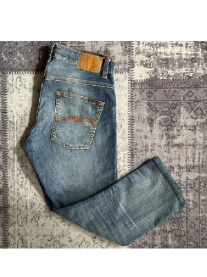 Blå jeans från Nudie Jeans - Säljer ett par klassiska blå jeans från Nudie Jeans med snygga slitningar och kontrastsömmar. Jeansen har rak passform, fem fickor och en ikonisk läderpatch bak i midjan. Perfekta för en avslappnad och trendig look.