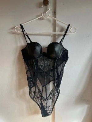Svart spetsbody med skinnlook-detaljer - Snygg svart body med bygel och vadderade kupor i skinnimitation. Genomskinlig mesh och spets med blommönster, smala axelband och markerade sömmar framtill. Perfekt för dig som vill ha något extra till din garderob. Byst: A eller B