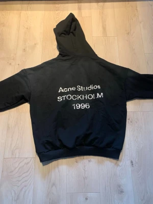 Acne studios hoodie - Svart hoodie från Acne Studios med tryck på ryggen: 'Acne Studios STOCKHOLM 1996'. Klassisk modell med huva och ribbade muddar. Perfekt för en avslappnad och trendig streetwear-look.