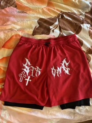 Röda shorts med tryck/streetwear/y2k/graphic shorts - Väldigt sköna röda shorts med vita grafiska tryck och kors på framsidan. Shortsen har elastisk midja med snörning och svarta innershorts. Baksidan har en ficka med dragkedja. Perfekt storlek för att ha hörlurar där. Perfekta för träning eller chill under sommaren.    O.B.S  trycket lossnar lite på ett ställe annars är allt på plats.