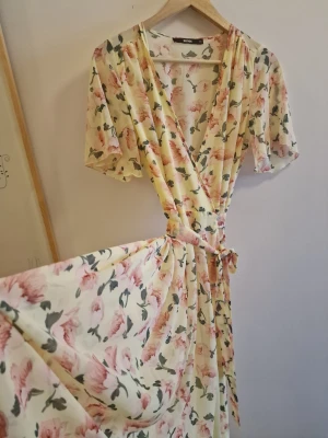 Blommig omlottklänning i pastell - Superfin lång omlottklänning från BIKBOK i ljus pastellgul med rosa blommor och gröna blad. Klänningen har korta ärmar, v-ringning och knyt i midjan för en snygg passform. Perfekt för sommarens alla tillfällen!