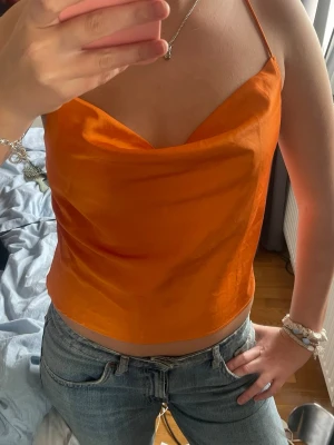 Orange linnetopp med tunna axelband - Snygg orange linnetopp med tunna axelband som korsas i ryggen. Toppen har en lös passform och ett mjukt, glansigt material som ger en lyxig känsla. Perfekt för varma dagar och somriga outfits. Super snygg och helt ny med prislappen kvar❣️