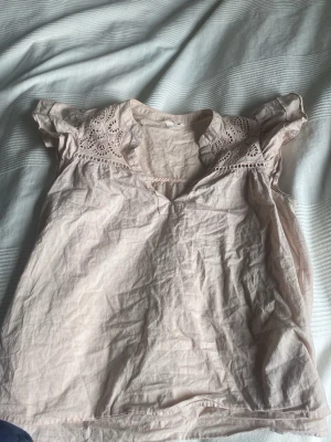 Ljusrosa blus med spetsdetaljer - Söt ljusrosa blus med korta ärmar och fina spetsdetaljer vid axlarna och halsringningen. Blusen har en lätt v-ringning och är tillverkad i ett tunt, mjukt tyg som ger en luftig känsla. Perfekt för en avslappnad och feminin look.