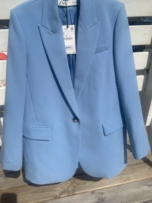Ljusblå kavaj från Zara - Aldrig använd. Nypris 859:-, nu 500:-. Snygg ljusblå kavaj från Zara med klassisk passform, spetsiga slag och enkelknäppning. Kavajen har två fickor med lock framtill och en bröstficka. Perfekt för att lyfta din outfit med en pop av färg.