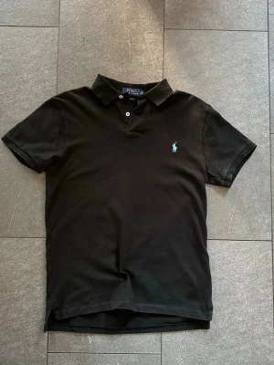 Svart pikétröja från Polo Ralph Lauren - Svart pikétröja från Polo Ralph Lauren i slim fit-modell. Tröjan har klassisk krage, knappar vid halsen och den ikoniska broderade loggan i ljusblått på bröstet. Tillverkad i bomull för en skön känsla. M men passar som S