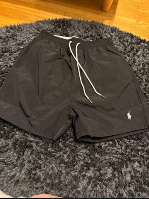 Svarta badbyxor från Polo Ralph Lauren - Säljer ett par stilrena svarta badbyxor från Polo Ralph Lauren med vitt broderat logotyp på benet och vita dragsnören i midjan. Klassisk passform och snabbtorkande material, perfekta för stranden eller poolen.