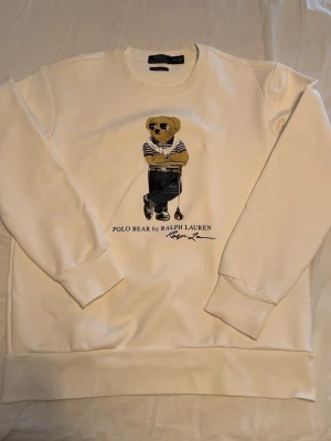 Vit sweatshirt Polo Bear Ralph Lauren - Vit sweatshirt från Ralph Lauren med ikoniskt Polo Bear-motiv framtill. Tröjan har rund halsringning, långa ärmar och ribbade muddar. Materialet är mjukt och bekvämt, perfekt för en avslappnad stil.