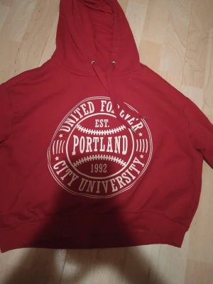 Röd hoodie Portland City University - Snygg röd hoodie med stor vit tryck på bröstet där det står 'United Forever Portland City University 1992'. Klassisk modell med huva och dragsko. Mjuk och skön bomullsmix, perfekt för chill dagar eller när du vill ha en sportig look.