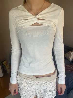 Vit ribbad långärmad topp twistad - Trendig vit ribbad topp med lång ärm och twistad detalj över bröstet som skapar en snygg cut-out. Toppen har en kroppsnära passform och är tillverkad i mjukt stretchigt material. Perfekt för dig som vill ha en unik och stilren look.