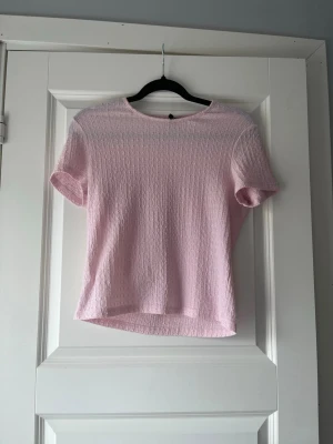 Ljusrosa kortärmad topp - Söt ljusrosa topp med korta ärmar och rund halsringning. Toppen har en diskret strukturerad yta som ger en extra detalj till looken. Perfekt att matcha med jeans eller kjol för en fräsch stil.