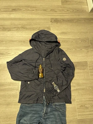 Marinblå Moncler vindjacka med huva - Snygg svart vindjacka från Moncler med huva och dragkedja framtill. Jackan har Moncler-logga på vänster ärm och praktiska fickor med knapp. Tillverkad i lätt och smidigt material som passar perfekt för blåsiga dagar.