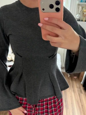 Mörkgrå peplumtop med utsvängda ärmar - Snygg mörkgrå peplumtop med långärmade, utsvängda ärmar och rund halsringning. Toppen har en figurnära passform upptill och markerad midja som ger en feminin siluett. Perfekt att styla med jeans eller kjol.