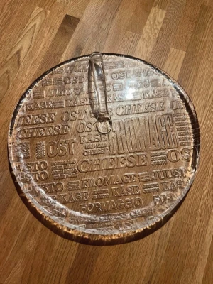 Ostbricka vintage i glas m handtag - Säljer en rund ostbricka i transparent glas med handtag. Brickan har relieftext med ordet ost på flera språk, som cheese, fromage och kase. Perfekt för servering av ost och snacks. Snygg detalj till köket eller middagsbjudningen. 30cm i diameter. 