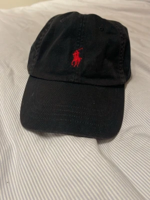 Svart keps från Polo Ralph Lauren - Svart keps i bomull från Polo Ralph Lauren med klassisk böjd skärm. Framtill finns den ikoniska röda polospelaren broderad och baktill står det 'POLO' i rött. Justerbart spänne bak för perfekt passform.