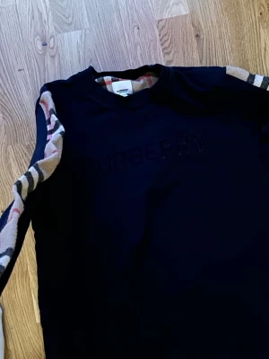 Svart Burberry sweatshirt med rutiga detaljer - Svart sweatshirt från Burberry med klassiska beige, röda och svarta rutiga detaljer längs ärmarna. Tröjan har rund halsringning och Burberry-loggan broderad framtill. Perfekt för dig som gillar stilrena plagg med exklusiv känsla. Använt länge. Storlek M