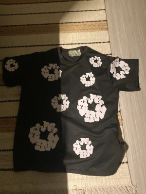 Svart t-shirt från Denim Tears - Säljer en svart t-shirt från Denim Tears med unika vita blommönster över hela tröjan. T-shirten har rund halsringning och korta ärmar. Perfekt för dig som vill sticka ut med en cool och modern stil.