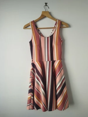 Randig klänning från Colloseum XS - Säljer en ärmlös klänning från Colloseum i storlek XS. Klänningen har ett färgglatt randigt mönster i rosa, vinrött, vitt, gult och svart. Modellen har en markerad midja och utställd kjol, tillverkad i ett mjukt och stretchigt material.