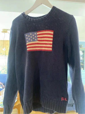 Mörkblå stickad tröja med USA-flagga - Stickad tröja i mörkblått med en stor amerikansk flagga i rött, vitt och blått på bröstet. Tröjan har långa ärmar och ribbade muddar vid ärmslut och nederkant. Perfekt för dig som gillar klassisk preppy stil. 