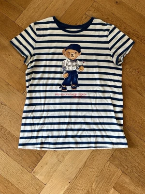 Randig Polo Bear t-shirt från Ralph Lauren - Vit och marinblå randig t-shirt från Ralph Lauren med Polo Bear-tryck framtill. Klassisk rund halsringning och korta ärmar. Tillverkad i mjuk bomull, perfekt för en avslappnad och cool stil.