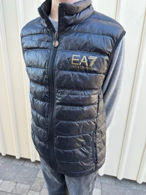 Svart dunväst från Emporio Armani EA7 - Snygg svart dunväst från Emporio Armani EA7 med guldfärgad logga på bröstet. Västen har hög krage, dragkedja framtill och quiltad design. Perfekt för lager-på-lager och ger en sportig look.