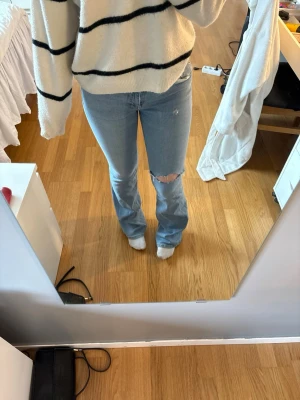 Ljusblå bootcut jeans med hål - Säljer ett par ljusblå bootcut jeans med snyggt slitage och hål på ena knät. Jeansen har klassisk femficksdesign och låg midja.