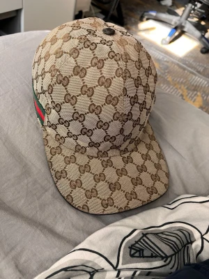 Gucci beige keps med GG-mönster - Säljer en beige keps från Gucci med det klassiska GG-monogrammet i brunt över hela kepsen. Kepsen har en böjd skärm och en grön-röd rand på sidan. Tillverkad i canvasmaterial som ger en exklusiv känsla och streetwear-vibe.