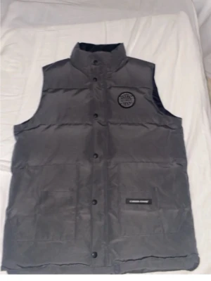 Grå dunväst från Canada Goose - Snygg grå dunväst från Canada Goose med hög krage och knäppning framtill. Västen har ett broderat svartvitt Canada Goose-märke på bröstet och en diskret logotyp på ena fickan. Perfekt för lager-på-lager och streetwear.