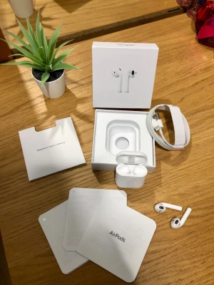 Apple AirPods trådlösa hörlurar - Säljer ett par vita Apple AirPods med laddningsetui och originalförpackning. Hörlurarna är trådlösa och har en stilren, minimalistisk design i plast. Allt som behövs för att komma igång ingår, inklusive laddkabel och manualer.