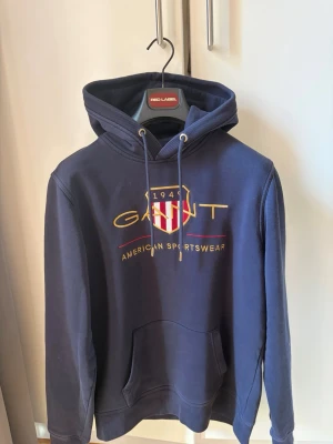 Gant Shield Hoodie - Snygg marinblå hoodie från Gant med broderad logga och texten 'American Sportswear' på bröstet. Klassisk huva med dragsko och stor magficka framtill. Tillverkad i mjukt bomullsmaterial och har ribbade muddar vid ärmslut och nederkant. 🎉OBS (Se bild 5 och 6) På varsin armbåge har materialet slitage därav pris! 🎉Inga returer accepteras då skick alltid beskrivs i beskrivning! 🎉För mer info skriv dm!