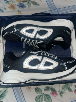 Svarta Dior B30 sneakers - Snygga svarta Dior B30 sneakers med stora vita CD-loggor på sidorna och vita detaljer. Ovandelen är i mesh och läder för en sportig vibe. Tjock vit sula och vita skosnören ger en clean kontrast. Perfekta för dig som vill sticka ut med exklusiva sneakers.