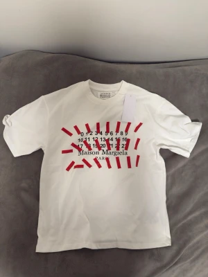 Vit Maison Margiela t-shirt med tryck - Vit t-shirt från Maison Margiela med ikoniskt siffertryck och röda grafiska detaljer framtill. Klassisk rund hals och korta ärmar. Perfekt för dig som gillar statement-plagg och modern design.