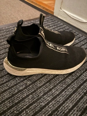 Svarta slip-on sneakers Michael Kors - Svarta slip-on sneakers från Michael Kors med stickad ovandel och tydlig logga på ovansidan. Skorna har vit sula med metallic-detalj vid hälen och elastisk passform för enkel påtagning. Perfekta för en trendig och sportig look.