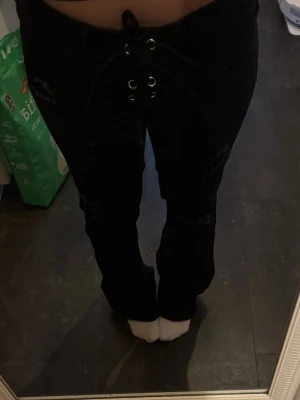 Svarta utsvängda jeans med snörning och hål - Snygga svarta jeans med utsvängda ben och cool snörning framtill i midjan. Jeansen har slitna detaljer på låren och knäna för en trendig look. Perfekta för dig som gillar en edgy och modern stil.