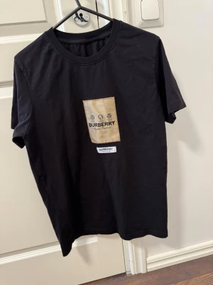 Svart Burberry t-shirt  - Svart t-shirt  med beige patch och logga på bröstet. Klassisk rund halsringning och korta ärmar. 