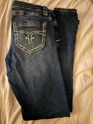 Mörkblå jeans med broderad bakficka - Snygga mörkblå jeans från Gina  med vit broderad detalj på bakfickan och slitningar. Hållen är npgot slitna men inget man lägger märke till. 