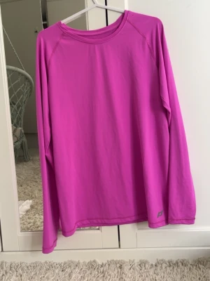 Rosa långärmad topp från Peak Performance - En enkel och sportig långärmad topp i stark rosa färg från Peak Performance. Tillverkad i ett mjukt och stretchigt syntetmaterial, perfekt för träning eller chill. Klassisk rund halsringning och diskret logga nere vid fållen.