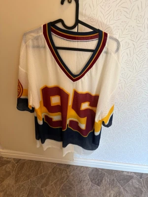 Vit mesh hockeytröja med nummer 95 - Säljer en vit mesh hockeytröja med stora siffran 95 i vinrött och gult framtill. Tröjan har breda ränder i gult och marinblått, v-ringad krage med vinröda och gula detaljer samt patch på ärmen. Perfekt för sport eller streetwear.