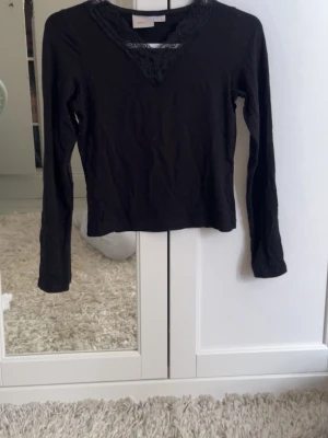 Svart långärmad topp med spets - Svart långärmad topp från Gina Tricot med v-ringning dekorerad med spetsdetaljer. Tillverkad i mjuk bomullsmix, perfekt för en stilren och enkel look. Passar till många olika outfits och är superenkel att matcha.